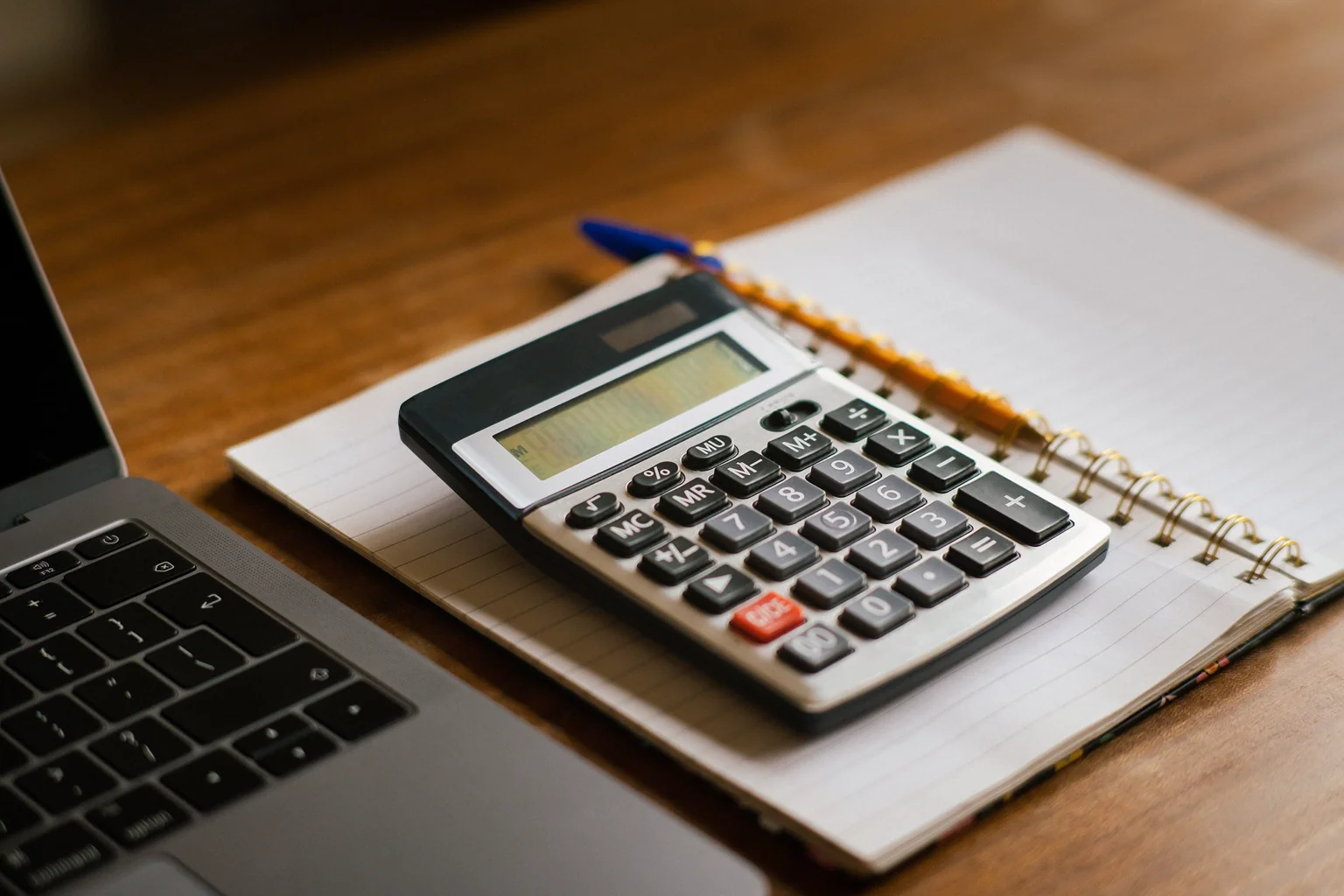 A calculator and notebook beside a laptop on a wooden desk.