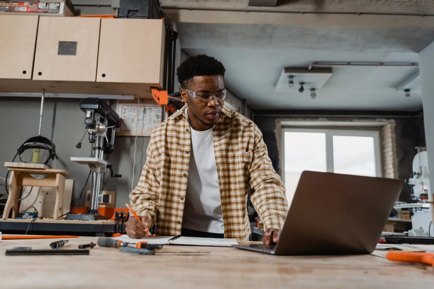 A tradesperson checking a laptop and project notes at a workshop bench.